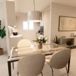 Zona pranzo contemporanea con tavolo in vetro, cucina bianca e piante decorative
